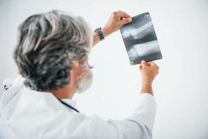 looks Bij röntgenfoto. senior mannetje dokter met grijs haar- en baard in wit jas is binnenshuis in kliniek foto