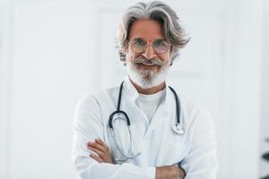 portret van senior mannetje dokter met grijs haar- en baard in wit jas staand binnenshuis in kliniek foto