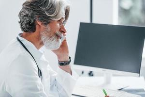 werken door computer tafel. senior mannetje dokter met grijs haar- en baard in wit jas is binnenshuis in kliniek foto