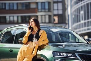 met smartphone. jong modieus vrouw in bordeaux gekleurde jas Bij dag met haar auto foto