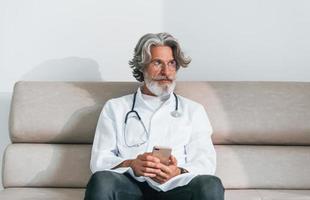 senior mannetje dokter met grijs haar- en baard in wit jas is binnenshuis in kliniek zit Aan sofa met telefoon in handen foto
