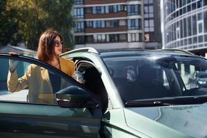 tegen bedrijf gebouw. jong modieus vrouw in bordeaux gekleurde jas Bij dag met haar auto foto