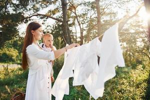 positief huisvrouw met blijft hangen wit kleren naar droog. jong moeder met haar weinig zoon is buitenshuis in de Woud. mooi zonneschijn foto