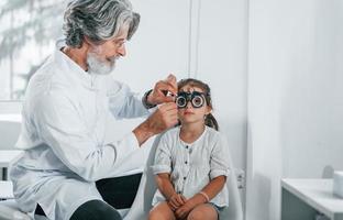 senior mannetje oogarts met grijs haar- en baard in wit jas is binnenshuis in kliniek testen zicht van weinig meisje foto