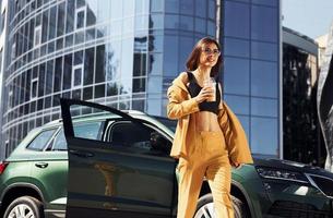 duurt breken met kop van koffie. jong modieus vrouw in bordeaux gekleurde jas Bij dag met haar auto foto