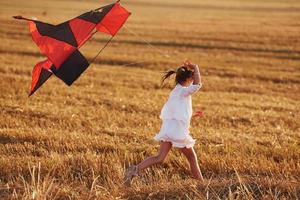 gelukkig weinig meisje rennen met rood vlieger buitenshuis Aan de veld- Bij zomertijd foto