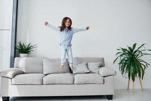 jumping Aan de bank. schattig weinig meisje binnenshuis Bij huis alleen. kind genieten van weekend foto