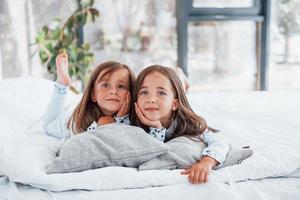 aan het liegen naar beneden Aan bed en op zoek Bij de camera. twee schattig weinig meisjes binnenshuis Bij huis samen. kinderen hebben pret foto