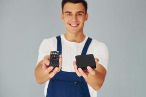 TV afgelegen controleur en smartphone. mannetje arbeider in blauw uniform staand binnen van studio tegen wit achtergrond foto