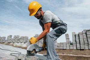 snijden door circulaire zaag. mannetje arbeider in geel gekleurde uniform hebben baan met bestrating foto