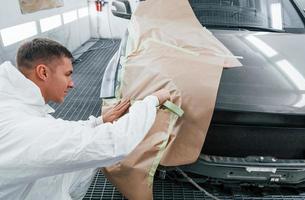 aan het bedekken auto door bruin lap. Kaukasisch auto- klusjesman in uniform werken in garage foto