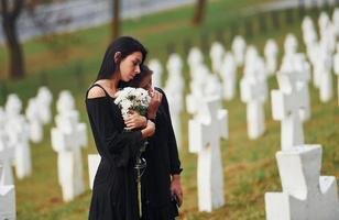 houdt bloemen. twee jong Dames in zwart kleren bezoekende begraafplaats met veel wit kruisen. opvatting van begrafenis en dood foto