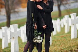 omarmen elk andere en huilen. twee jong Dames in zwart kleren bezoekende begraafplaats met veel wit kruisen. opvatting van begrafenis en dood foto