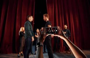 vrouw handen houdt telefoon en nemen afbeelding. groep van acteurs in donker gekleurde kleren Aan repetitie in de theater foto