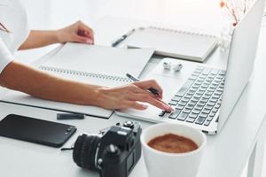 gebruik makend van laptop en kladblok. jong vrouw freelancer werken binnenshuis in de kantoor Bij dag foto