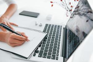 gebruik makend van laptop en kladblok. jong vrouw freelancer werken binnenshuis in de kantoor Bij dag foto