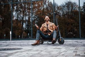 tegen gebouw en met elektrisch scooter. jong mannetje model- in modieus kleren is buitenshuis in de stad Bij dag foto