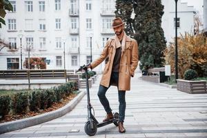 Aan de elektrisch scooter. jong mannetje model- in modieus kleren is buitenshuis in de stad Bij dag foto