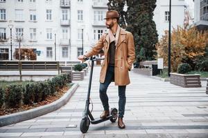 Aan de elektrisch scooter. jong mannetje model- in modieus kleren is buitenshuis in de stad Bij dag foto