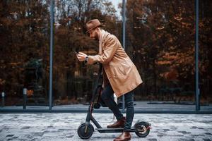 Aan de elektrisch scooter. jong mannetje model- in modieus kleren is buitenshuis in de stad Bij dag foto