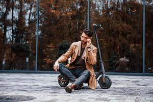 tegen gebouw en met elektrisch scooter. jong mannetje model- in modieus kleren is buitenshuis in de stad Bij dag foto