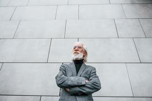 senior zakenman in formeel kleren, met grijs haar- en baard is buitenshuis staat tegen de muur foto