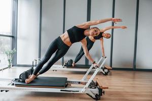 staand Aan Sportschool uitrusting en aan het doen strekt zich uit. twee Dames in sportief slijtage en met slank lichamen hebben geschiktheid yoga dag binnenshuis samen foto