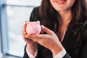 vrouw in zwart formeel kleren is binnenshuis in de modern kantoor houdt roze varkentje bank foto