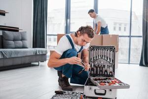 geval met apparatuur. twee jong verhuizers in blauw uniform werken binnenshuis in de kamer foto