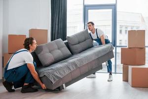 draag- zwaar bank. twee jong verhuizers in blauw uniform werken binnenshuis in de kamer foto