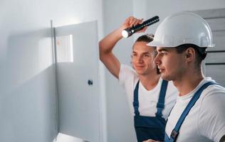 twee jong mannetje elektriciens werken binnenshuis samen. gebruik makend van zaklamp foto