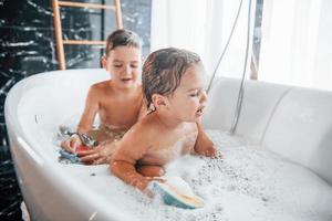 twee kinderen hebben pret en het wassen zich in de bad Bij huis. helpen elk andere foto