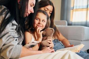 lezing boek samen. vrouw lesbienne paar met weinig dochter uitgeven tijd samen Bij huis foto