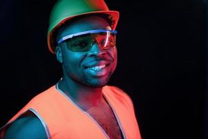 bouw arbeider in uniform en moeilijk hoed. futuristische neon verlichting. jong Afrikaanse Amerikaans Mens in de studio foto