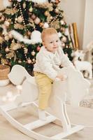 een blond meisje in een trui ritten een houten paard Bij huis tegen de achtergrond van de Kerstmis boom. schattig weinig meisje spelen Bij huis Aan de achtergrond van slingers. foto