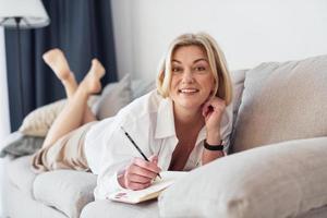 middelbare leeftijd vrouw in elegant kleren is Bij huis schrijven in kladblok foto