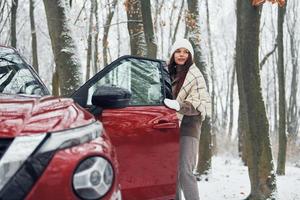 positief emoties. mooi jong vrouw is buitenshuis in de buurt haar rood auto- Bij winter tijd foto