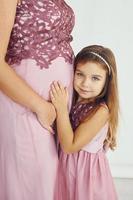 opvatting van anticipatie. zwanger moeder. vrouw in jurk staand met haar dochter in de studio met wit achtergrond foto