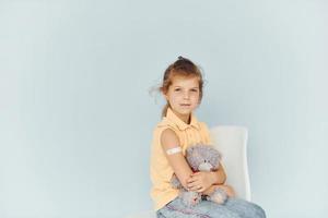 weinig meisje zittend Aan de stoel tegen wit achtergrond na vaccinatie foto