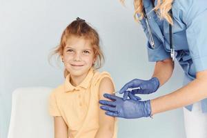 weinig meisje in geel shirt. dokter in uniform maken vaccinatie naar de geduldig foto