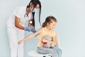 weinig meisje met teddy beer. dokter in uniform maken vaccinatie naar de geduldig foto