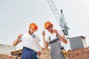 lezing plan. twee jong bouw arbeiders in uniformen is bezig Bij de onvoltooid gebouw foto