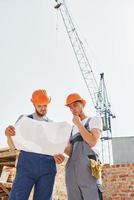 lezing plan. twee jong bouw arbeiders in uniformen is bezig Bij de onvoltooid gebouw foto