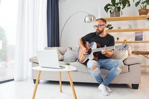 gebruik makend van laptop. Mens in gewoontjes kleren en met akoestisch gitaar is binnenshuis foto