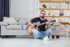 zittend in de buurt de bank. Mens in gewoontjes kleren en met akoestisch gitaar is binnenshuis foto