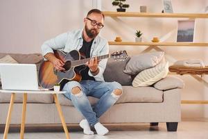 zittend Aan de bank. Mens in gewoontjes kleren en met akoestisch gitaar is binnenshuis foto