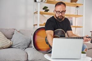 gebruik makend van laptop. Mens in gewoontjes kleren en met akoestisch gitaar is binnenshuis foto