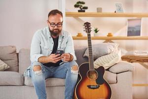 zittend Aan de bank. Mens in gewoontjes kleren en met akoestisch gitaar is binnenshuis foto