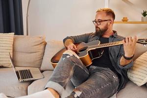 hebben mooi zo tijd beoefenen. Mens in gewoontjes kleren en met akoestisch gitaar is binnenshuis foto