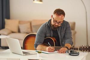 schrijven sommige ideeën. Mens in gewoontjes kleren en met akoestisch gitaar is binnenshuis foto